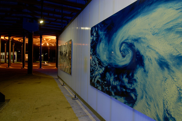 Zwei groformatige Fotos hängen an einer Ausstellungswand. Im Hintergrund ist ein Meteroit zu sehen