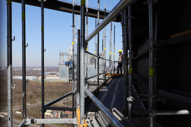 Ein Gerüstbauer auf dem Fassadengerüst des Gasometers unterhält sich mit einem Kollegen, der einen mobilen Aufzug bedient.